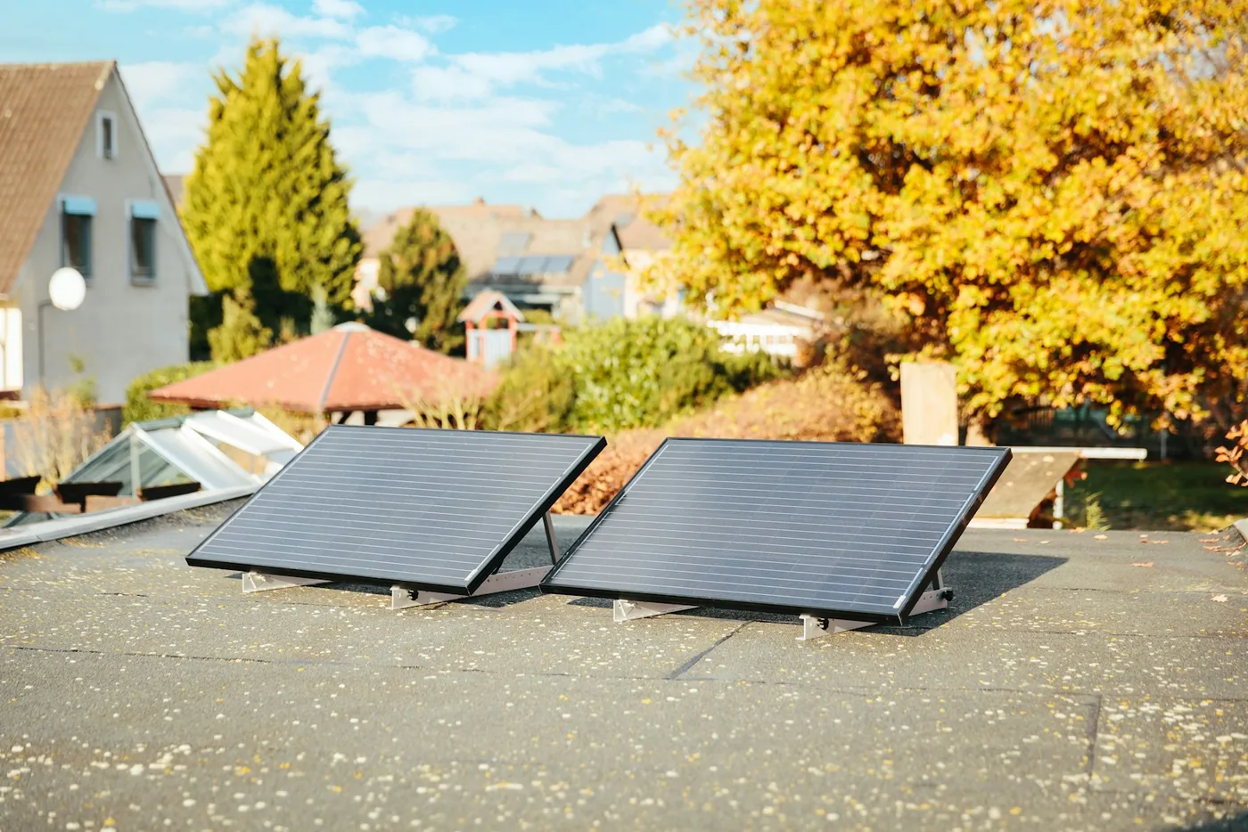 Zwei Solarpanels auf einem Flachdach, im Hintergrund herbstliche Bäume und ein Haus