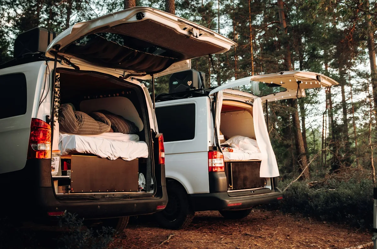 Zwei weiße Vans in einem Kiefernwald geparkt, Hecktüren offen, jeweils mit einem Bett über dem Batteriefach eingebaut
