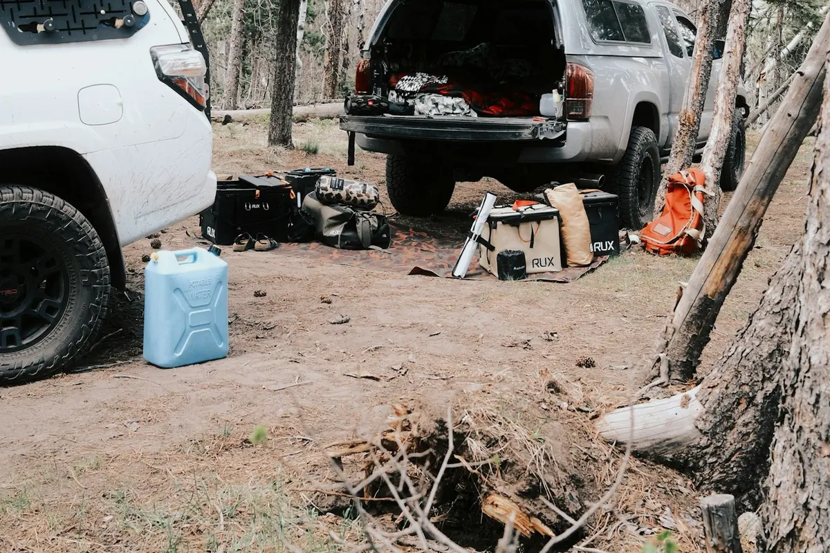 Two vehicles parked at a forest campsite with camping gear laid out on the ground and a blue water jerrycan