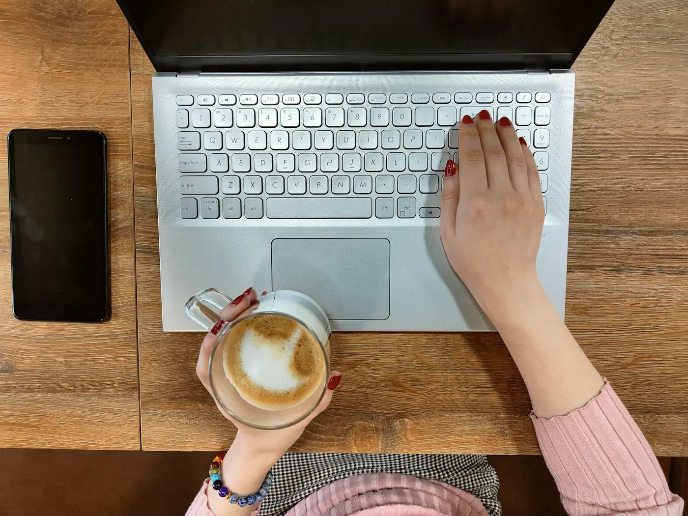 Top view of hands on a silver laptop keyboard with a latte in one hand and a phone on a wooden table