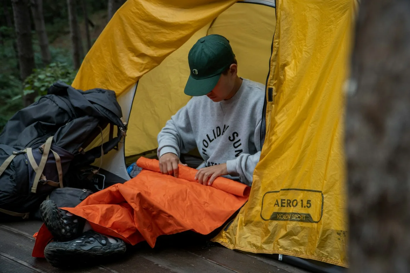 Una persona acampando con gorra organizando un objeto naranja en la puerta de una tienda amarilla, con una mochila al lado