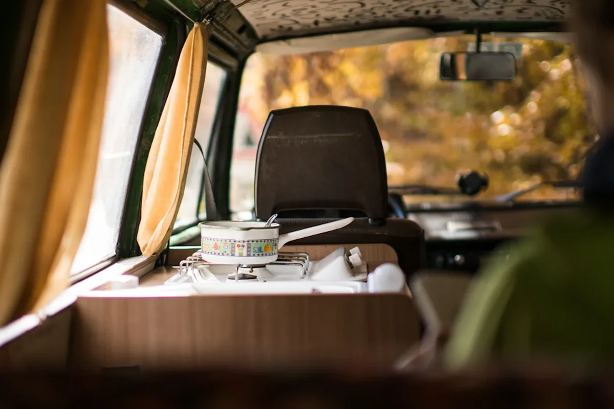 Een camperinterieur met een klein keukenblok, een kookpan en warm licht door het raam