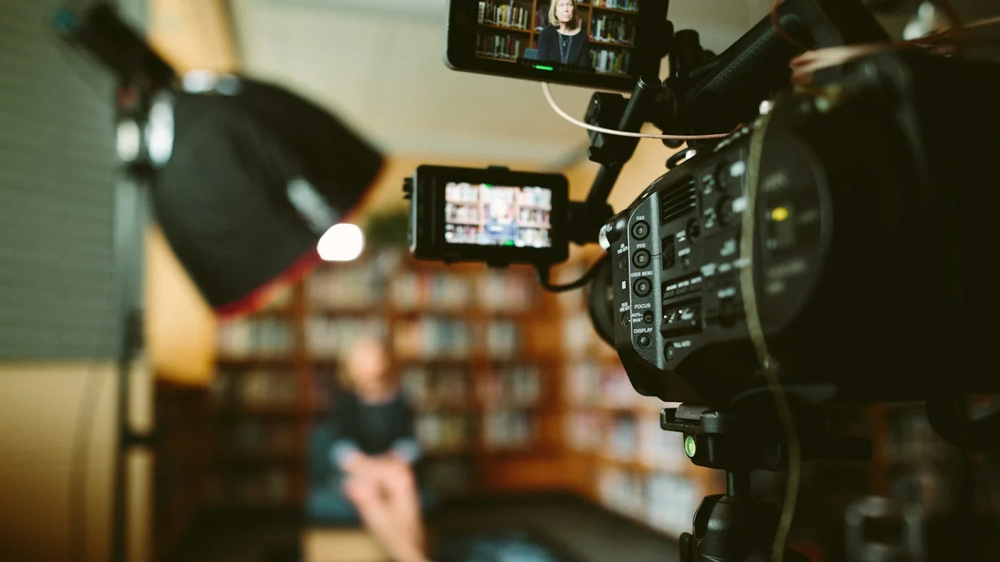 A video camera with a monitor filming a person seated in front of a bookshelf