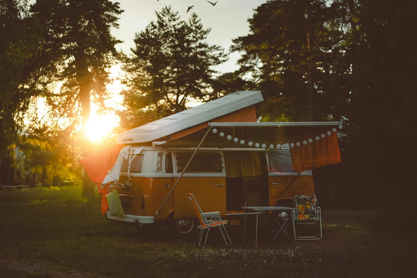 Oranger und weißer VW-Camper mit Hubdach auf einem Campingplatz zwischen Bäumen bei Sonnenuntergang mit Lichterketten