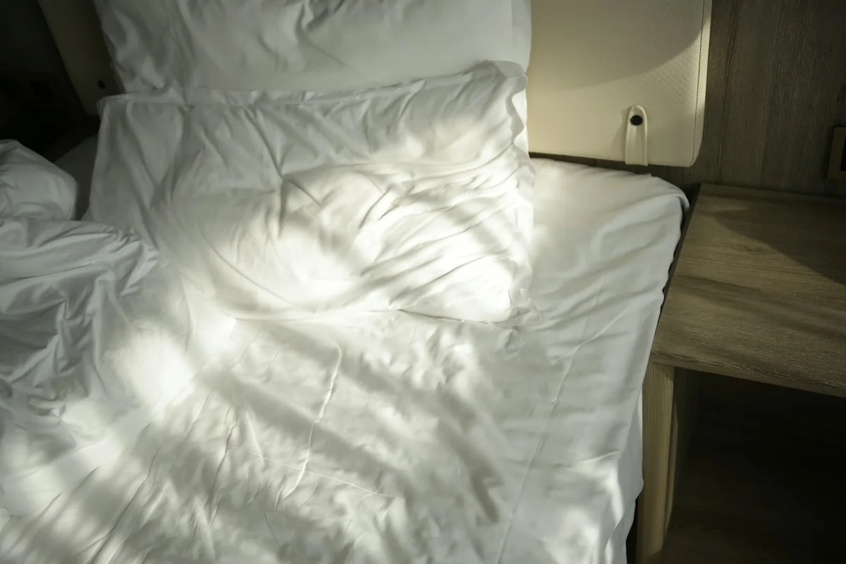 Rumpled white bed with pillows beside a wooden nightstand in soft light