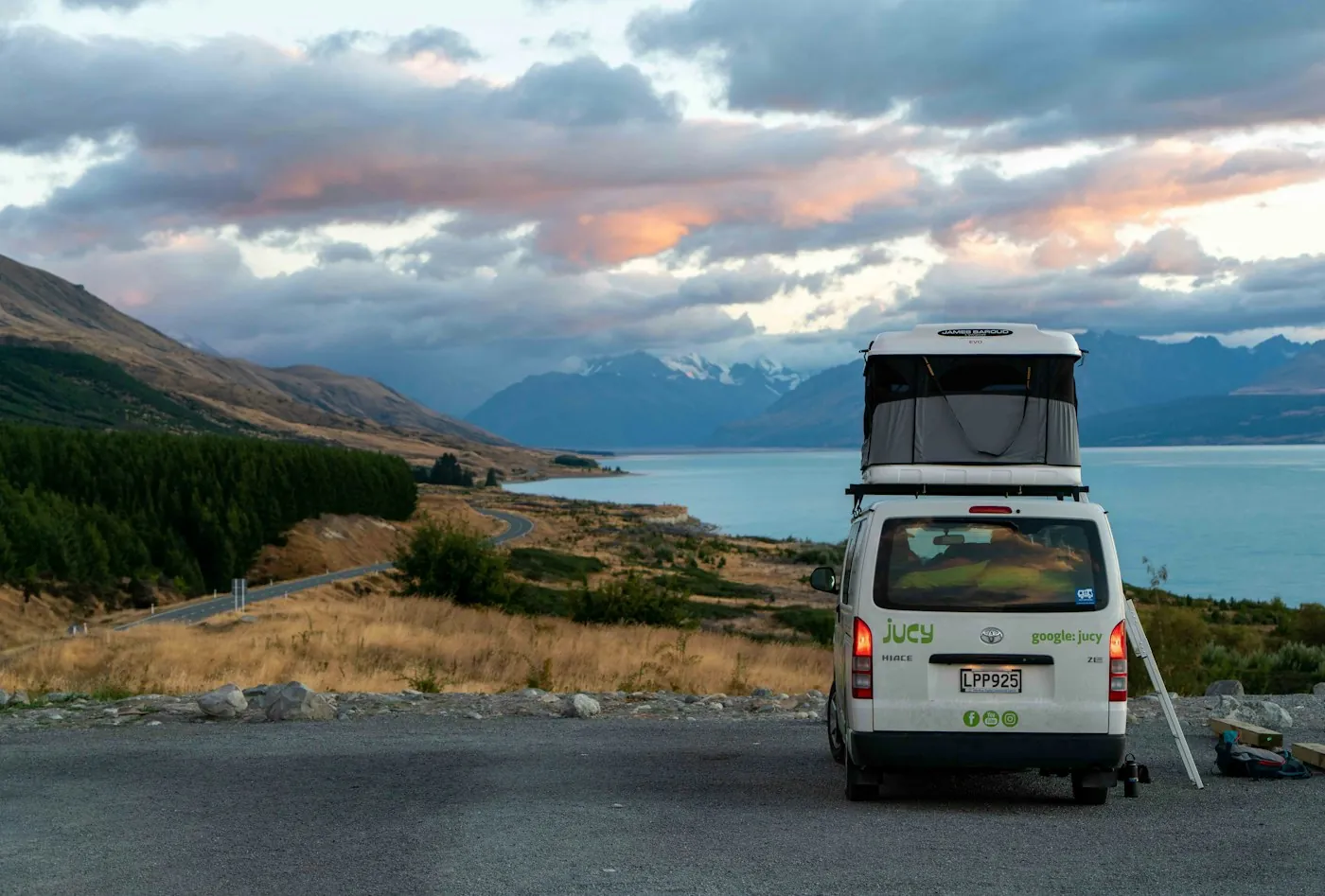 A white camper van parked on the road edge above a lake with mountains behind it