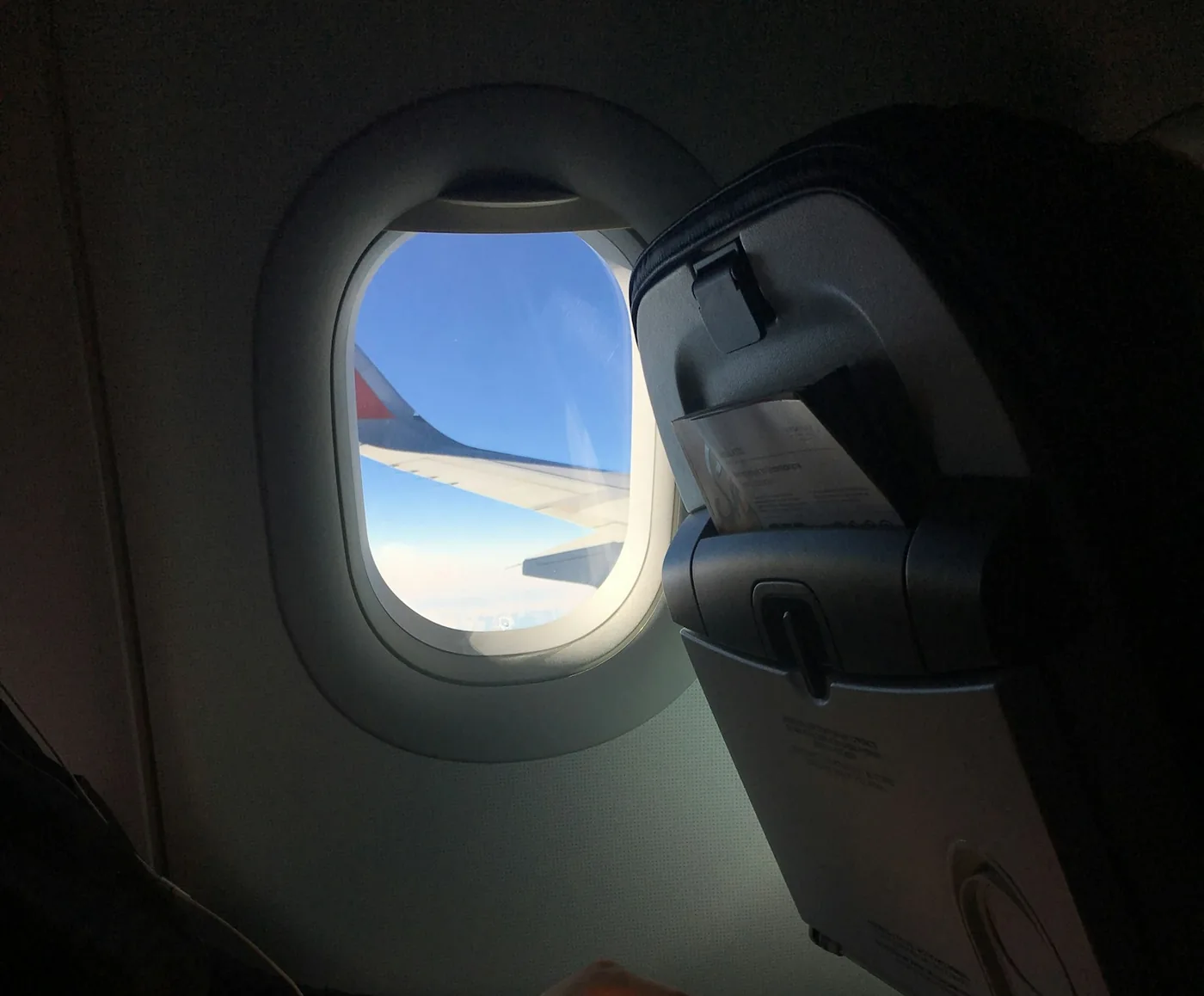 Airplane window with blue sky and a wing outside and a passenger's hands holding earbud cables in the dark cabin