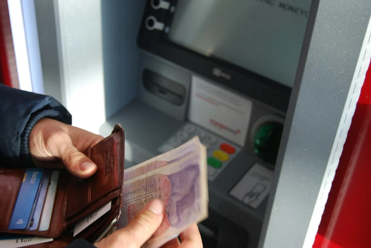 Hand holding a leather wallet and banknotes in front of an ATM while withdrawing cash abroad