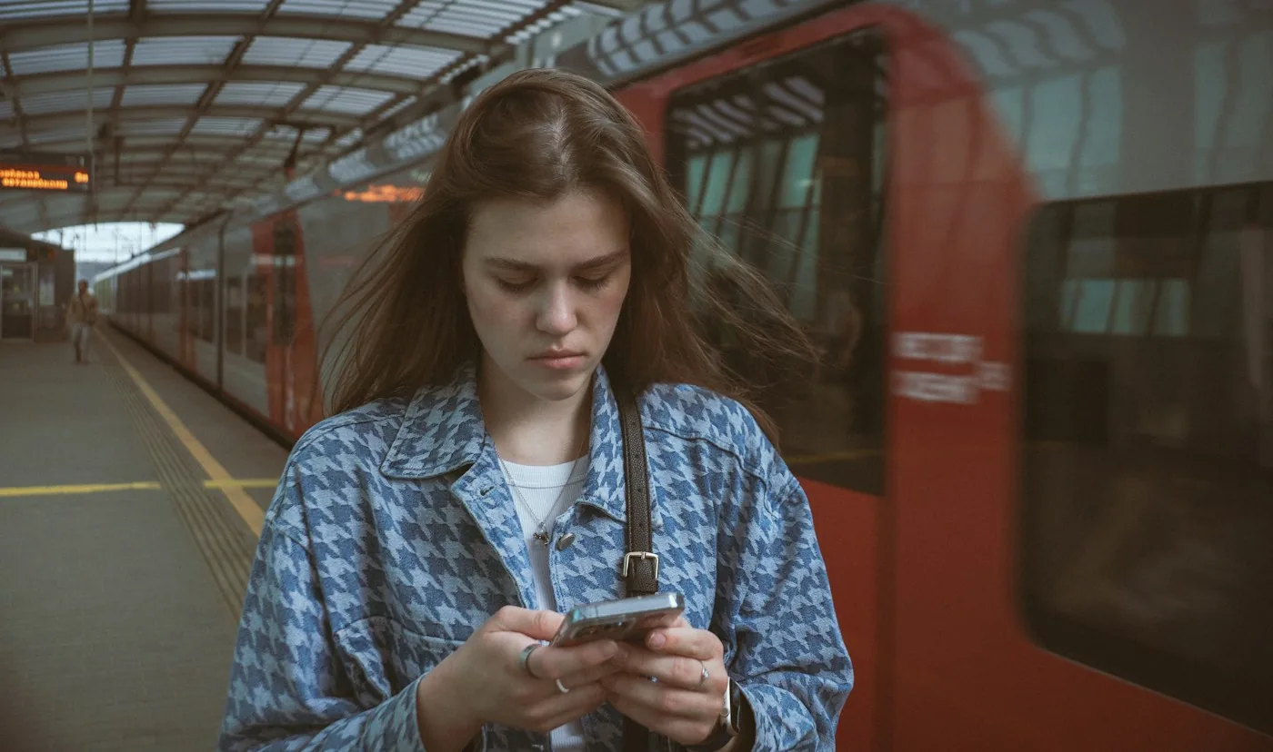 Woman in a patterned jacket checking her phone on a train platform with a red train behind her