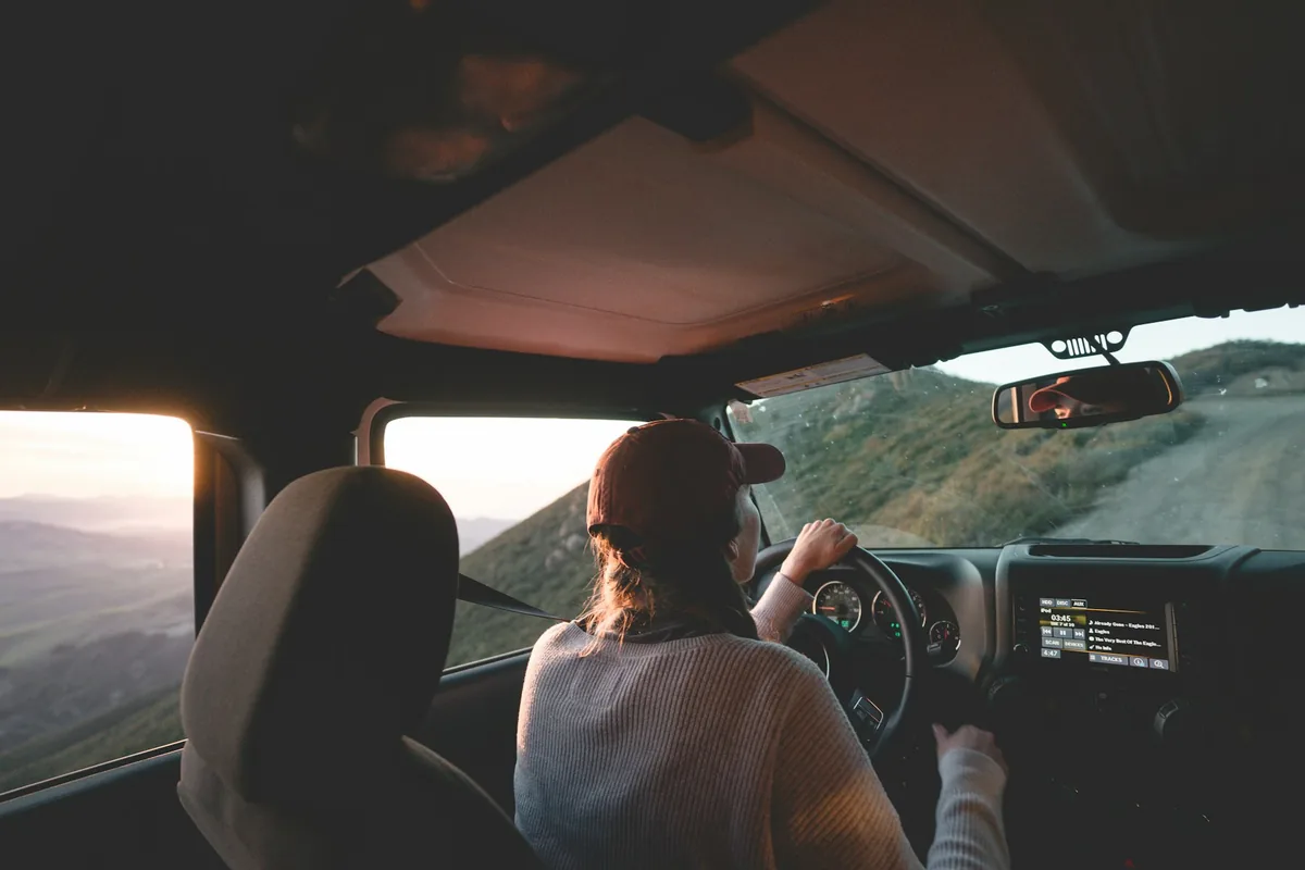 A woman driving on a mountain road at sunset, seen from the back seat