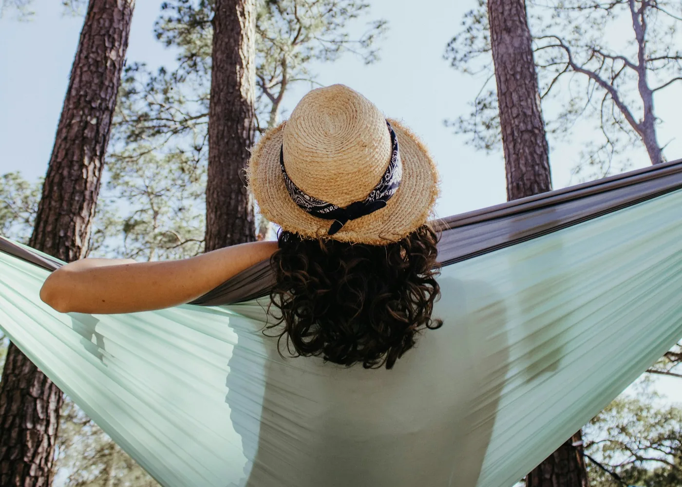 Woman in a straw hat lying in a light green hammock among tall pine trees