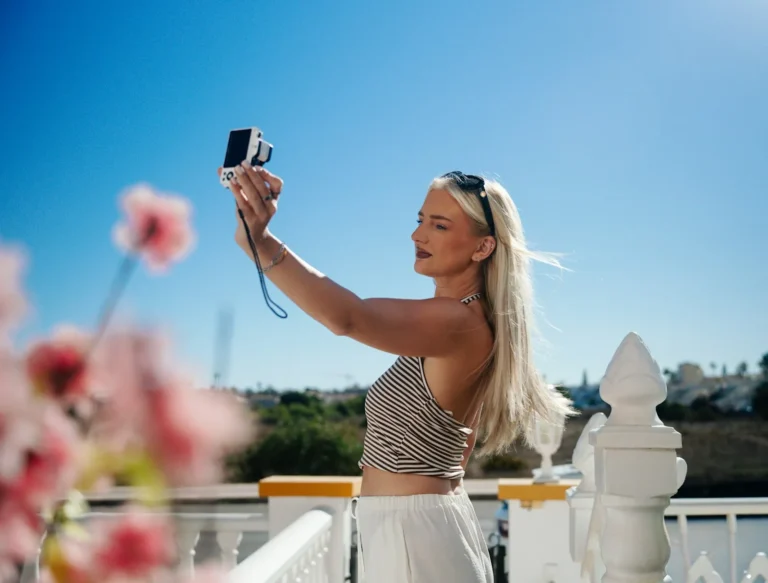 A woman holding a small camera at arm's length to film herself against a blue sky