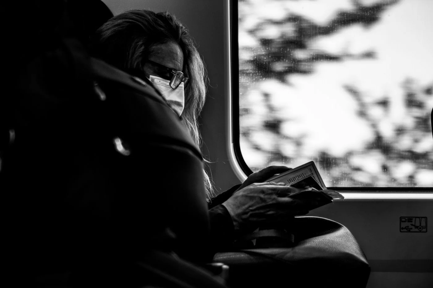 A woman in glasses reading a small book on a train, seen in black and white by the window
