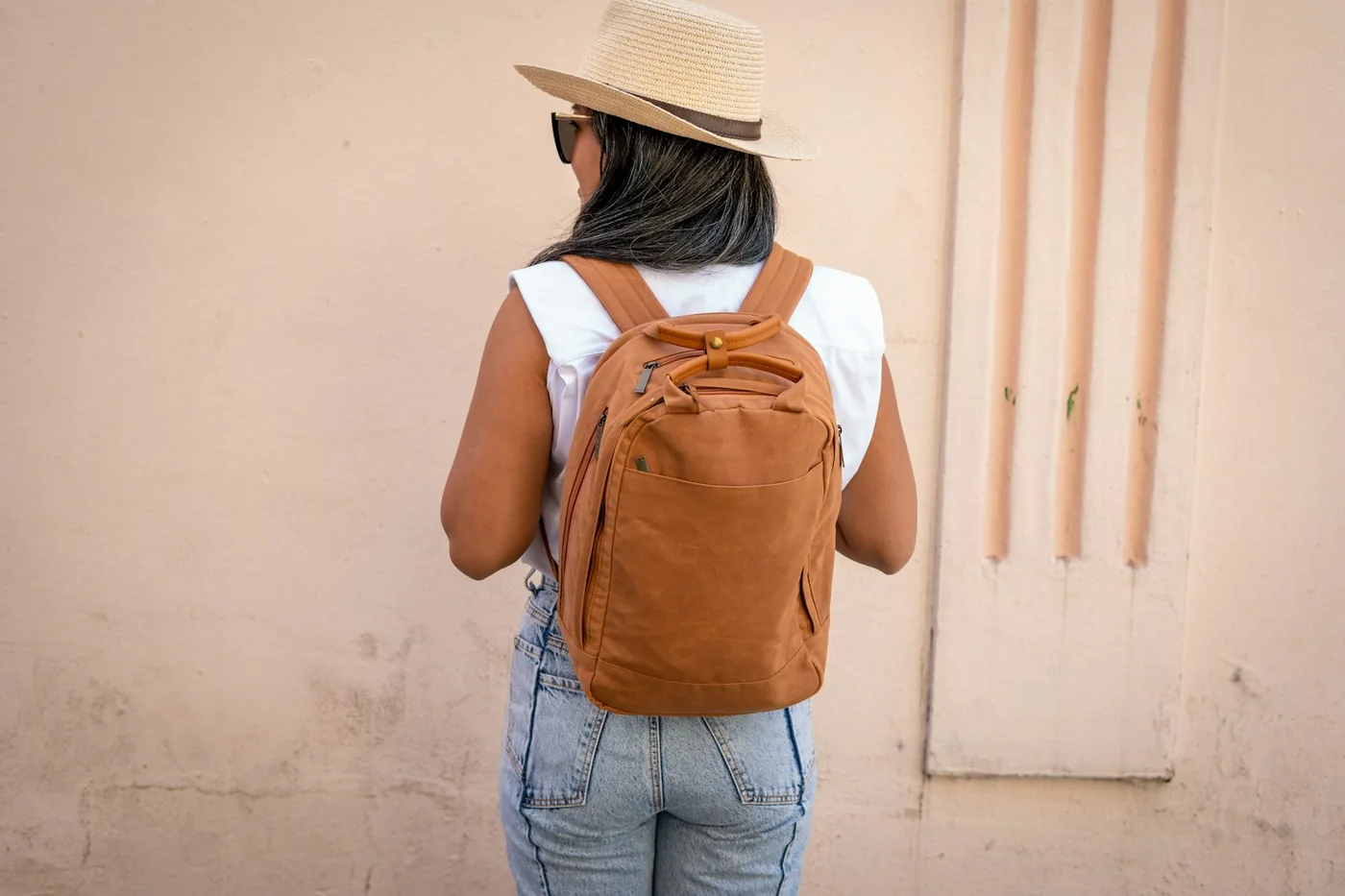 Una mujer con sombrero de paja llevando una mochila de día marrón, vista de espaldas, frente a una pared rosa