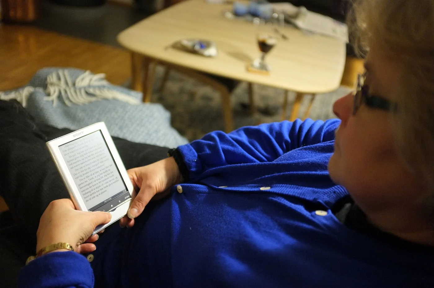 A woman in glasses and a blue cardigan lying back and reading on a white e-reader in a living room