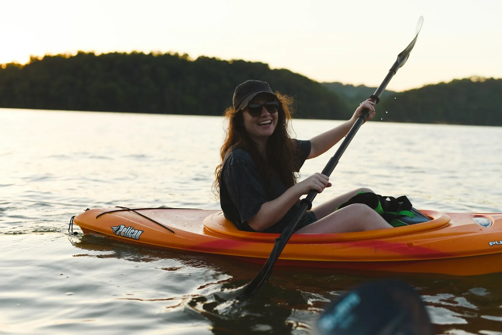 A smiling woman paddling an orange kayak on a calm lake at sunset