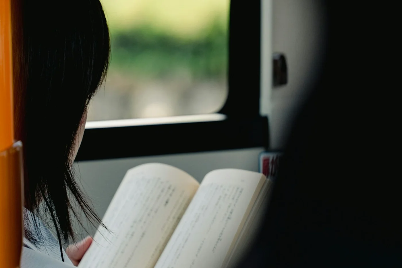 A woman with dark hair reading an open book by a bus window with trees outside