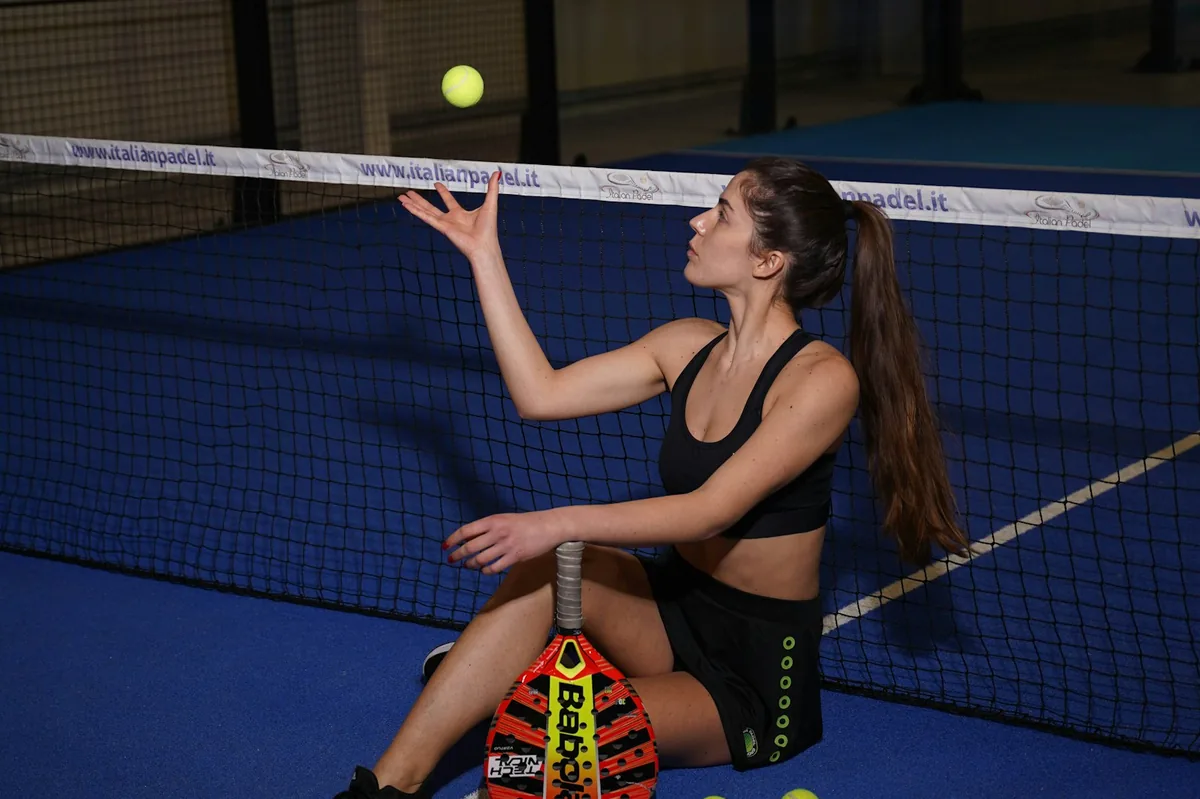 Frau wirft einen Ball über einem Padel-Platz hoch, im Hintergrund das Netz