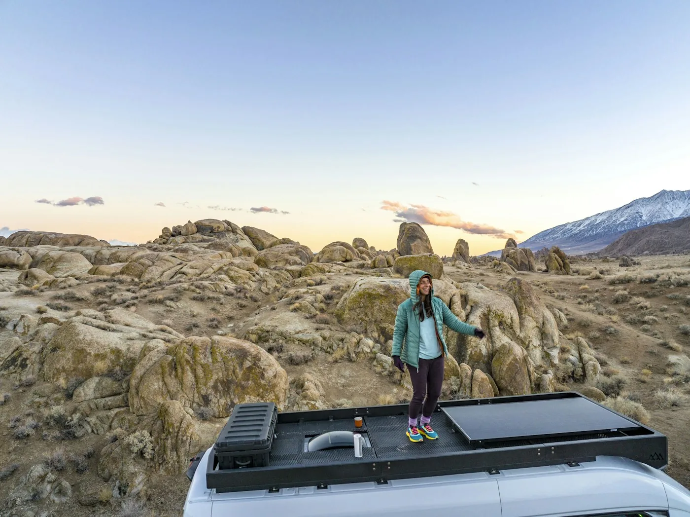 Woman in a teal jacket standing on a van roof with a solar panel in a rocky desert at dusk
