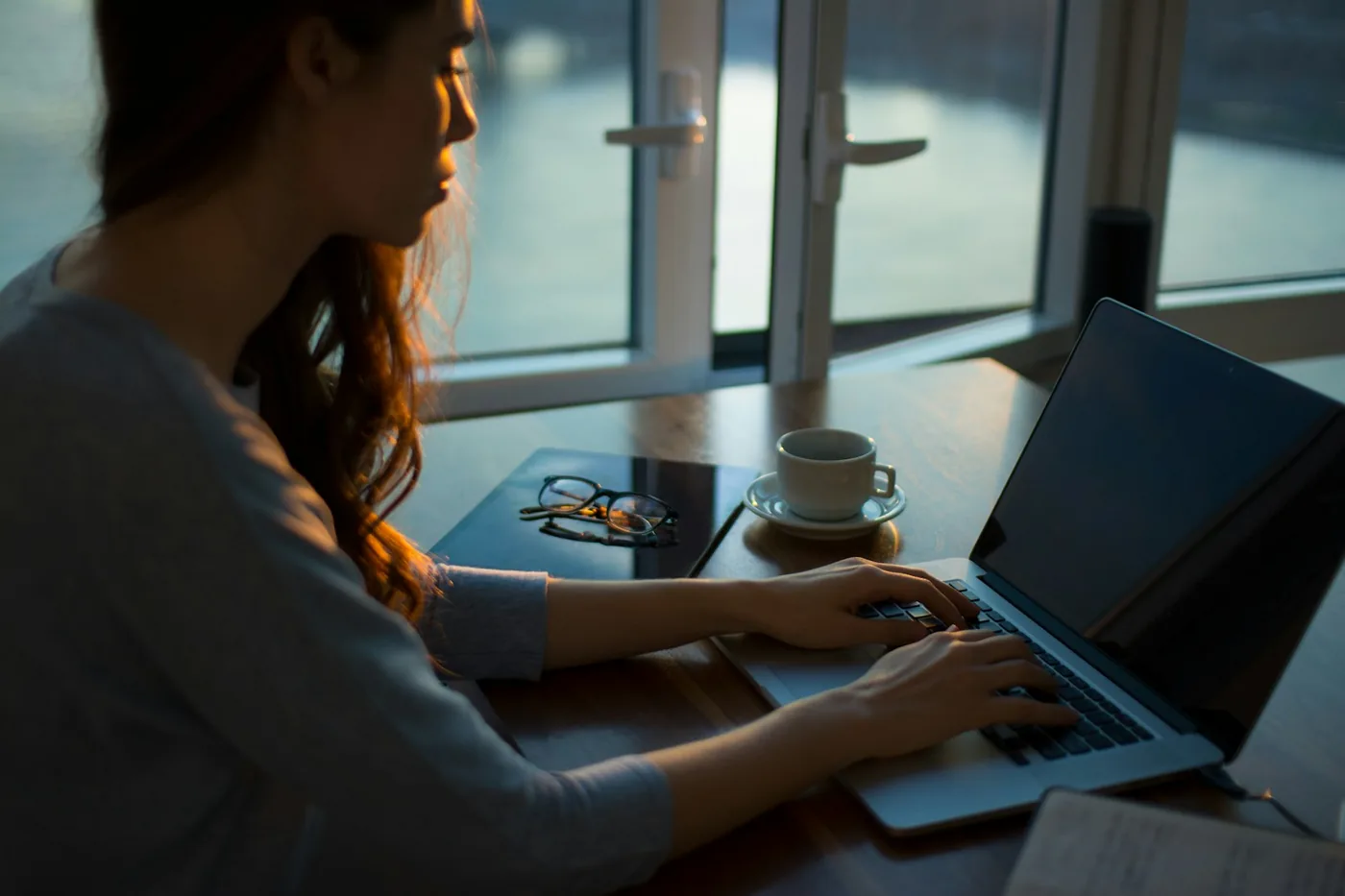A woman typing on a laptop at a table next to a coffee cup and a window