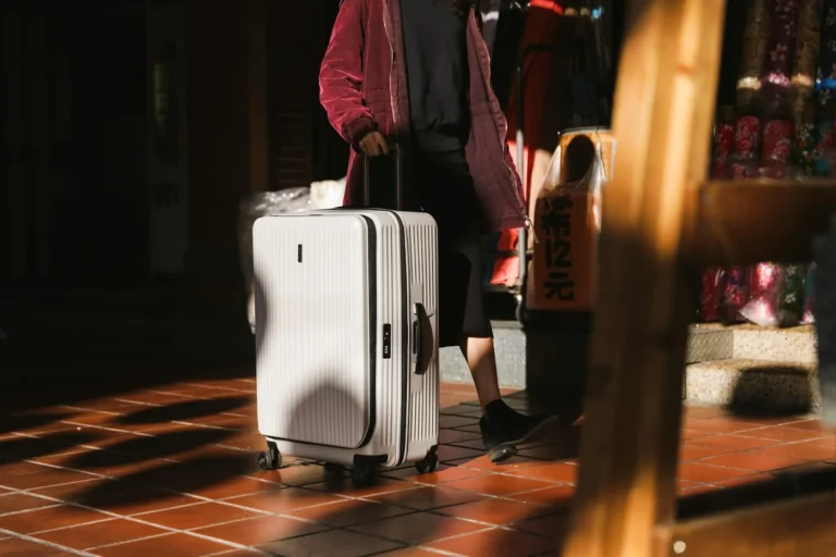 Woman in a pink jacket walking with a white hardside suitcase across a tiled floor in sunlight