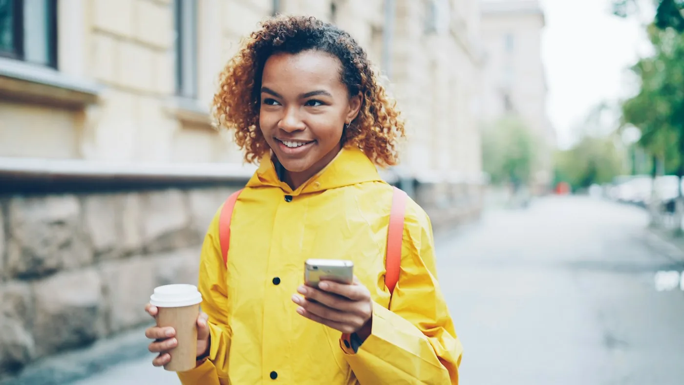 Woman in a yellow raincoat holding a coffee and checking her phone on a city street