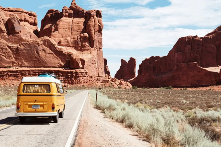 A yellow VW van driving on a desert road between red rocks