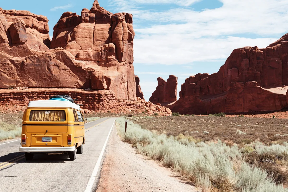 A yellow VW van driving on a desert road between red rocks